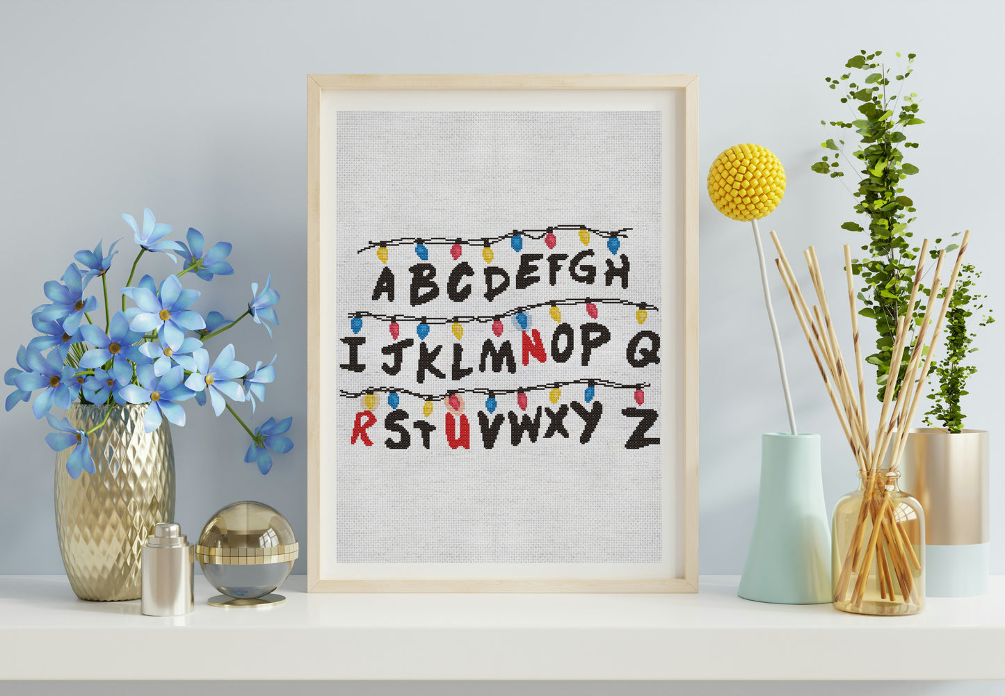 Framed alphabet print with decorative lights on a shelf with flowers and plants.