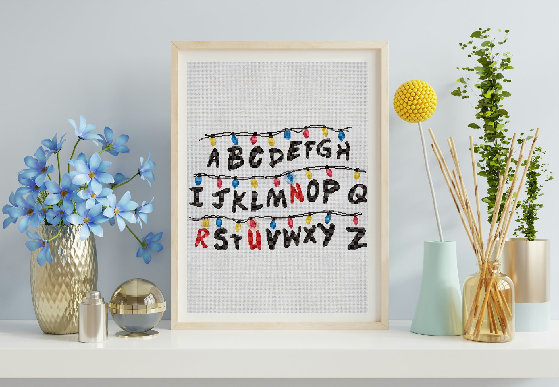 Framed alphabet print with decorative lights on a shelf with flowers and plants.