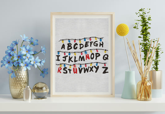Framed alphabet print with decorative lights on a shelf with flowers and plants.