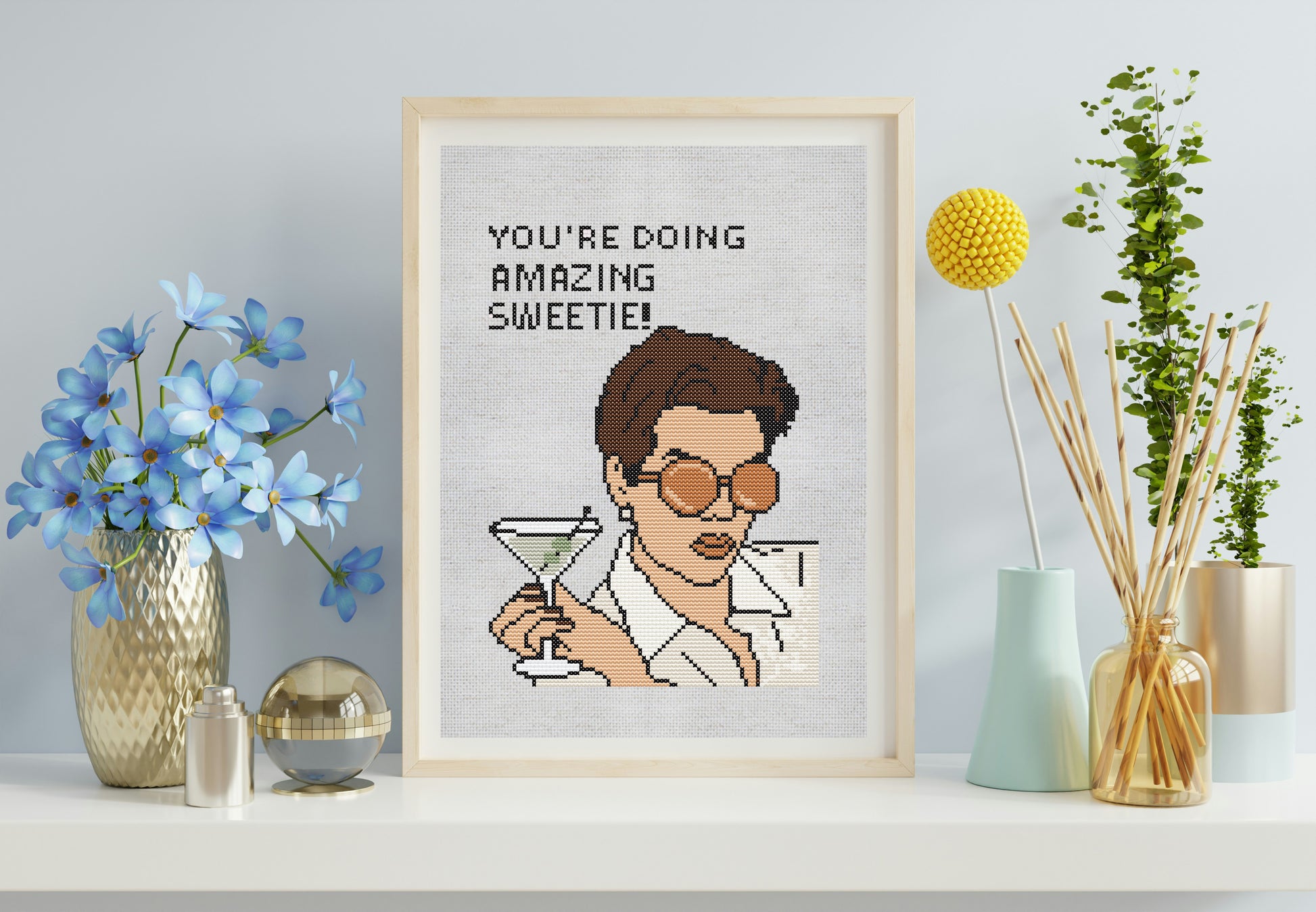 Framed cross-stitch art of a character holding a martini glass with text, placed on a shelf with decorative items.