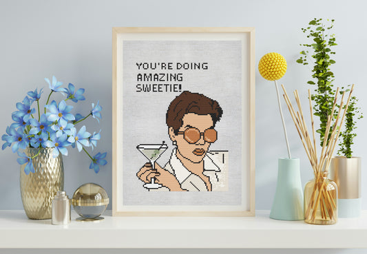 Framed cross-stitch art of a character holding a martini glass with text, placed on a shelf with decorative items.