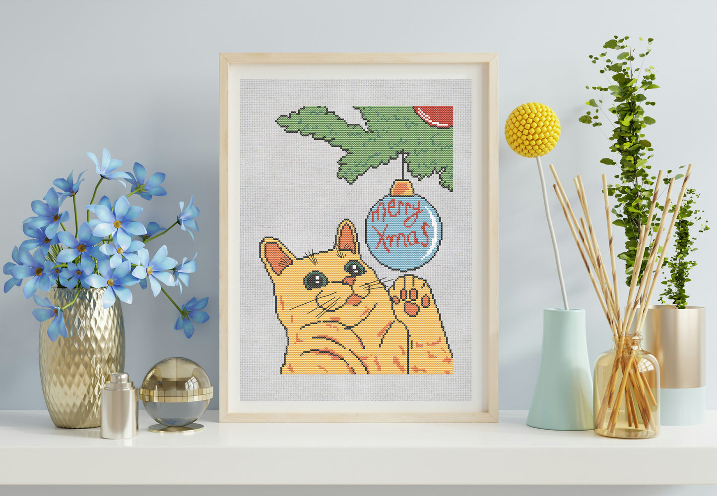 Framed cross-stitch of a cat with a Christmas ornament, surrounded by decorative items on a shelf.