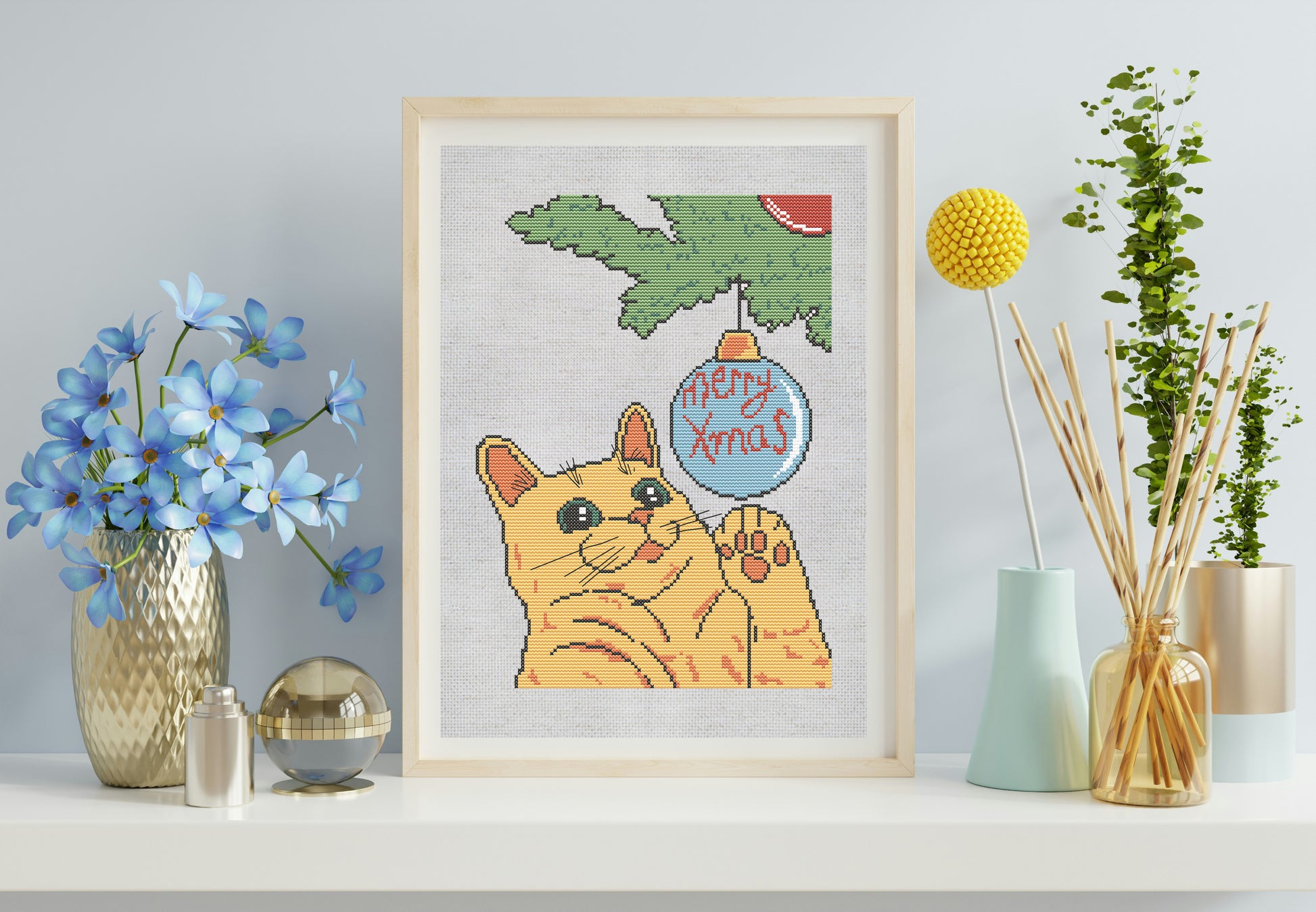 Framed cross-stitch of a cat with a Christmas ornament, surrounded by decorative items on a shelf.