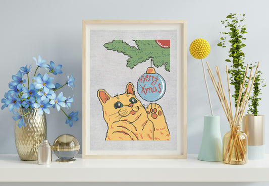 Framed cross-stitch of a cat with a Christmas ornament, surrounded by decorative items on a shelf.