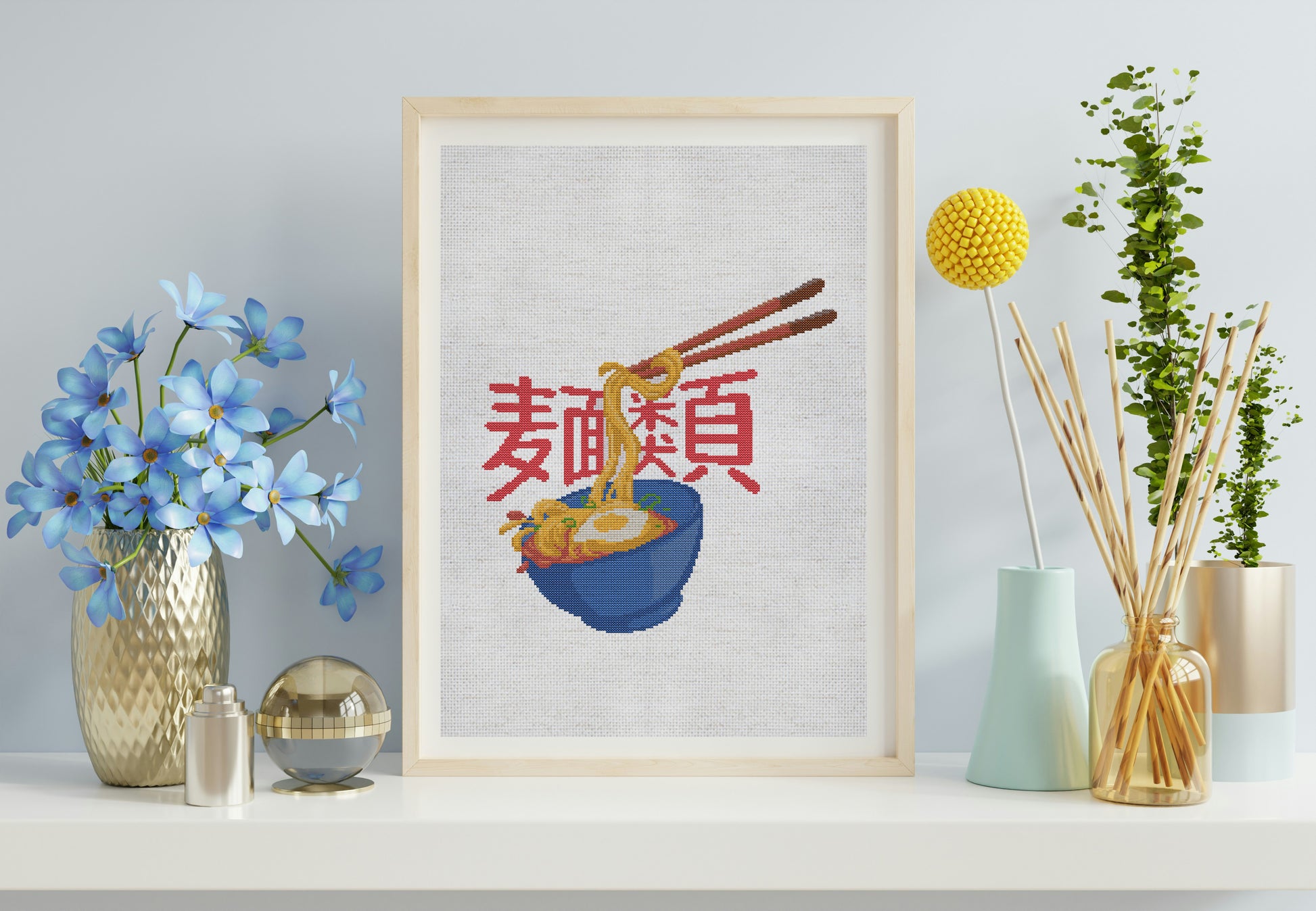 Framed cross-stitch art of a bowl of noodles with chopsticks on a shelf with decorative items.