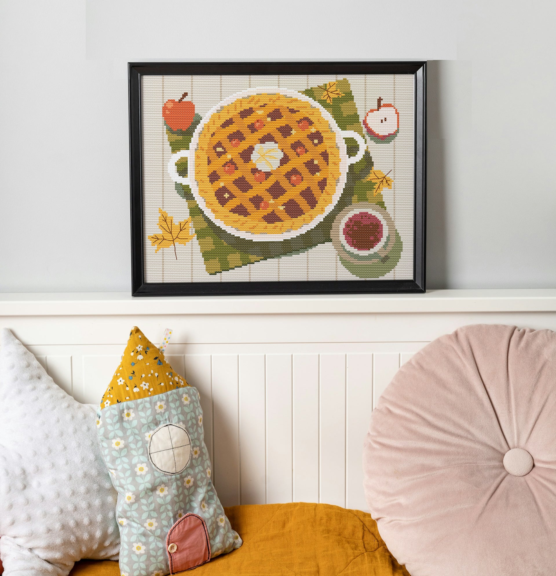 Framed cross-stitch art of a waffle with fruit on a wall above a sofa with decorative pillows.