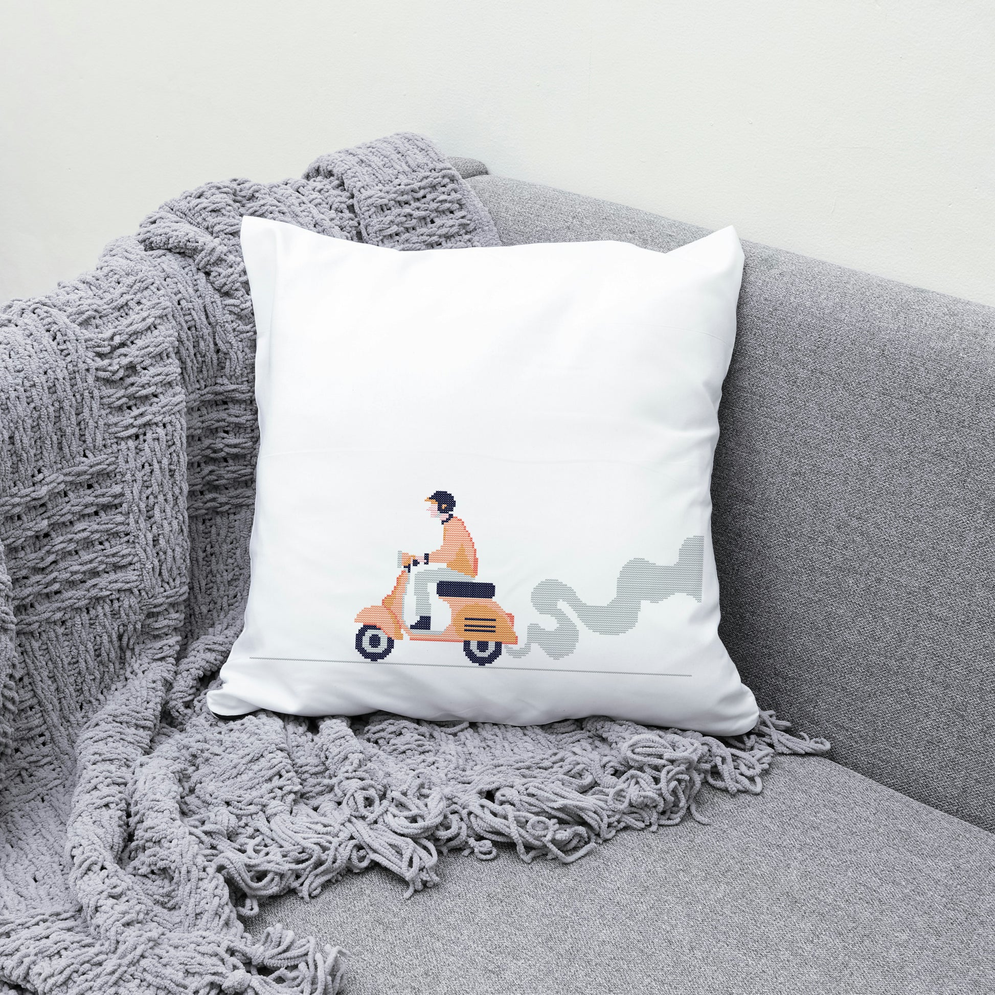 White pillow with a design of a person on a scooter on a gray couch with a textured blanket.