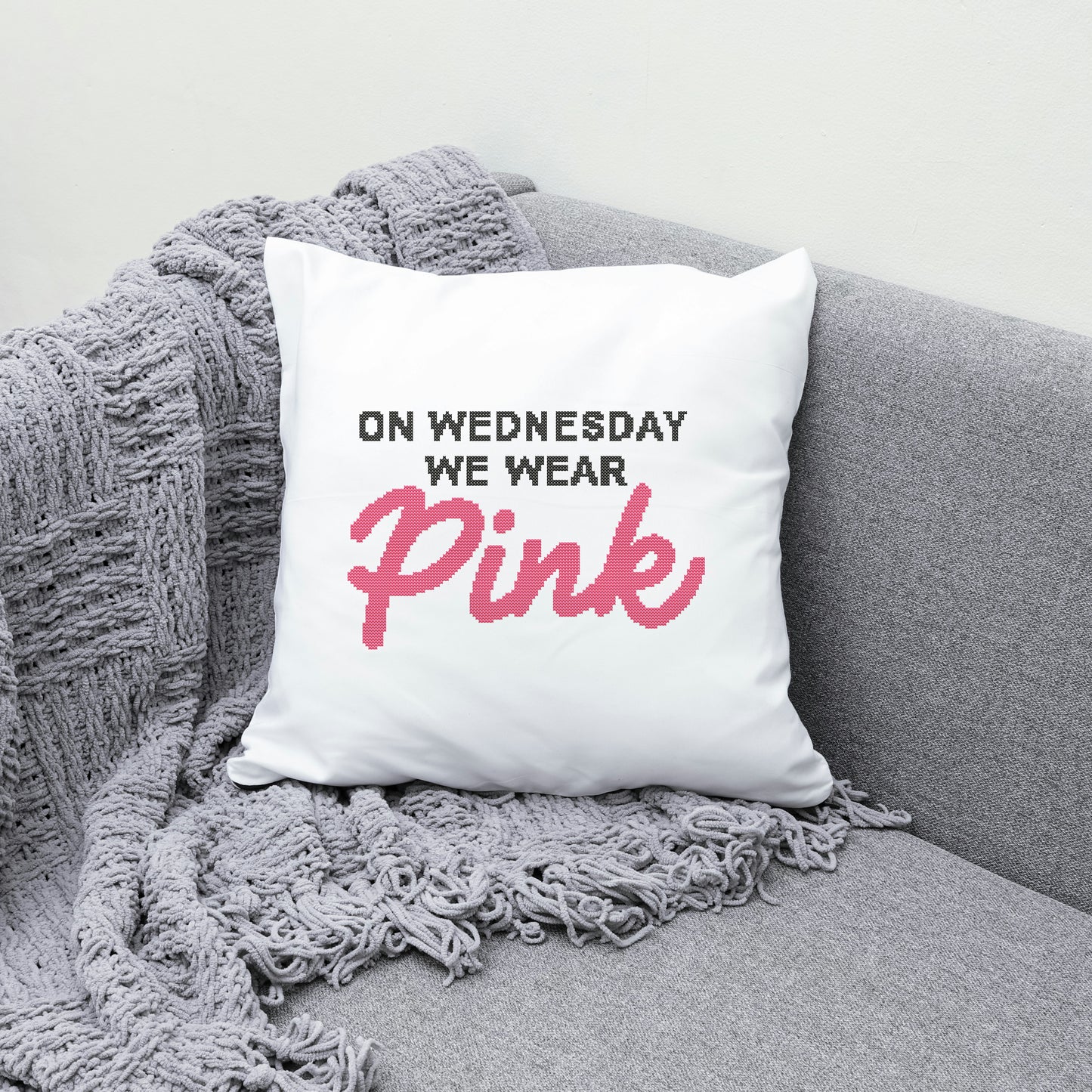 White pillow with text 'On Wednesday We Wear Pink' on a gray couch with a gray blanket.
