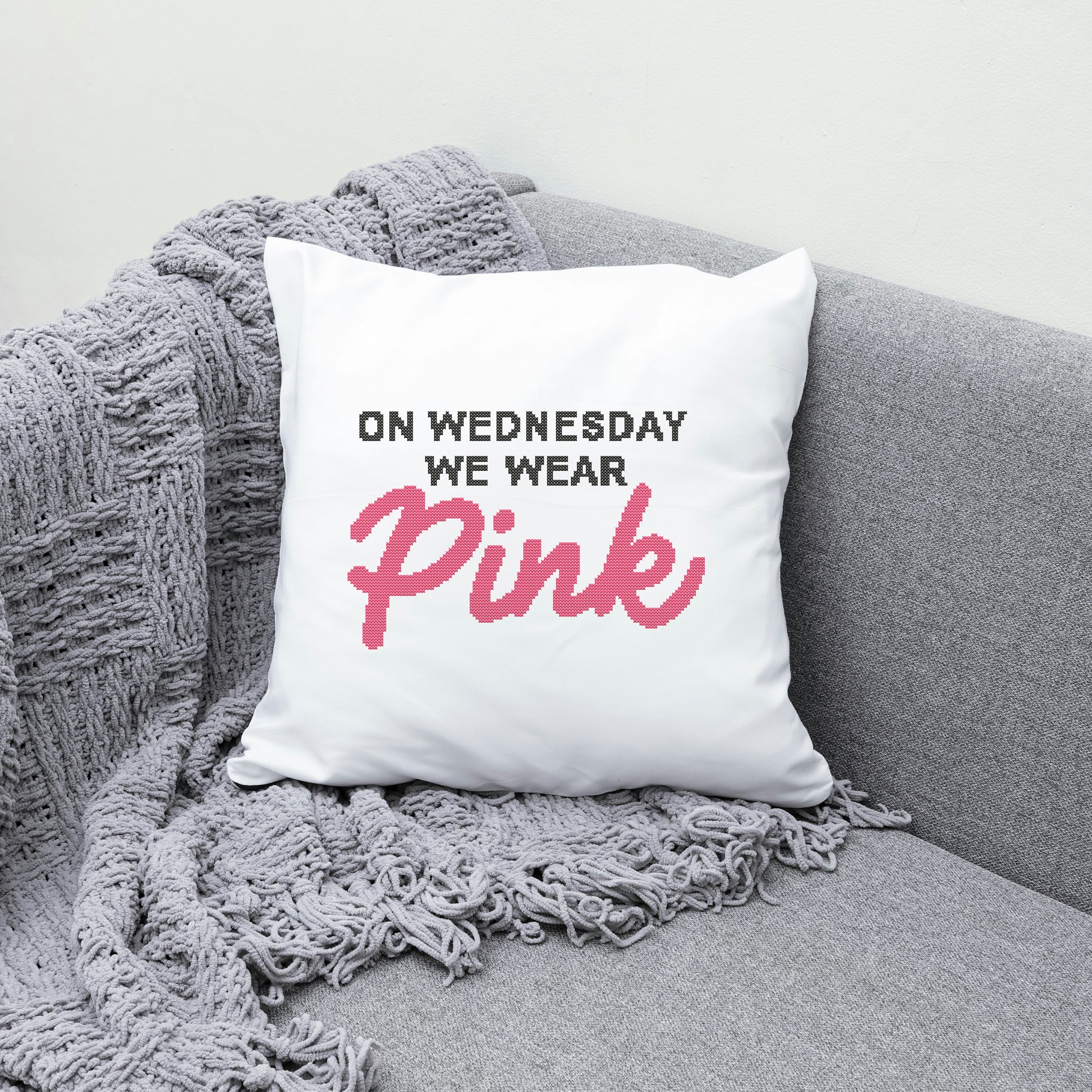 White pillow with text 'On Wednesday We Wear Pink' on a gray couch with a gray blanket.