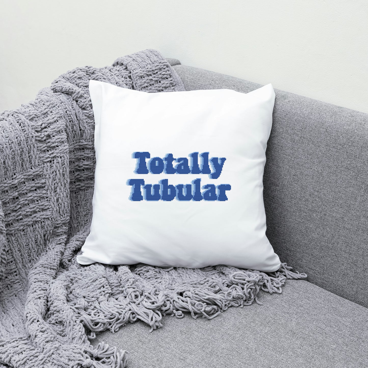 White pillow with 'Totally Tubular' text on a gray couch with a textured blanket.