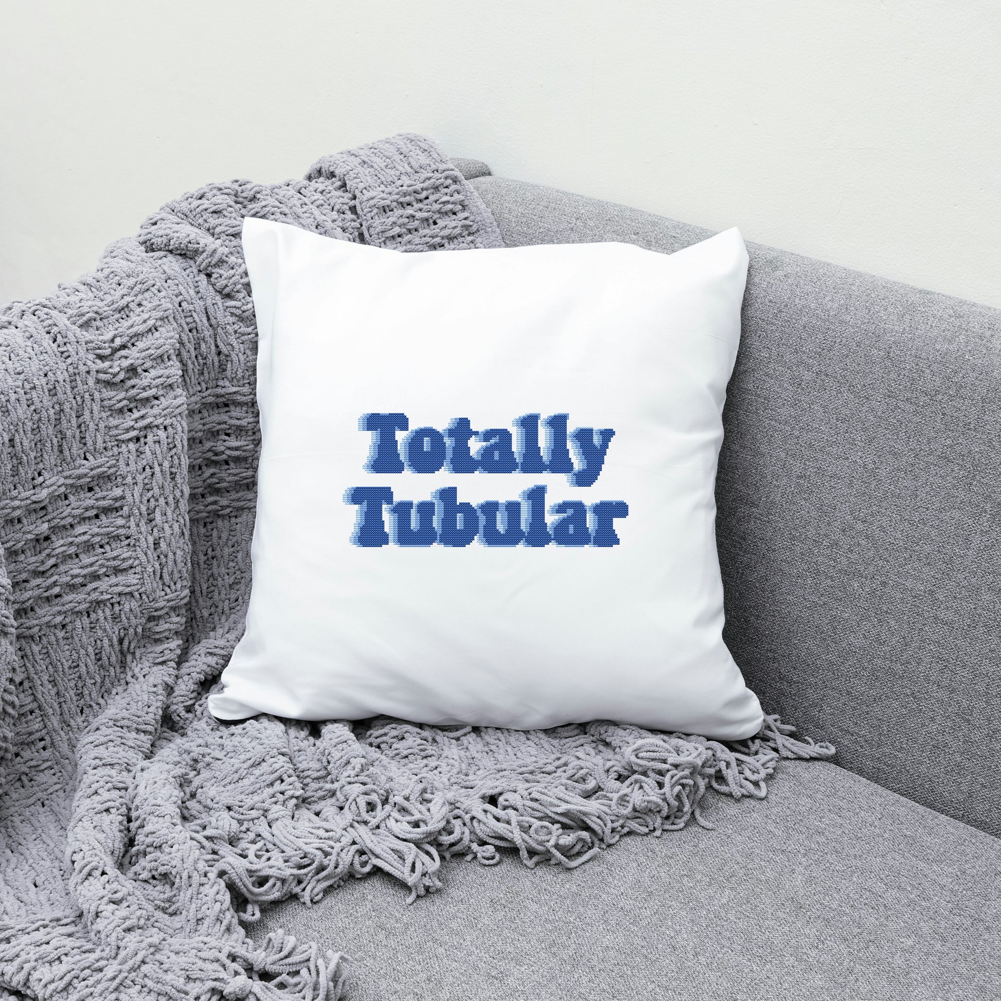White pillow with 'Totally Tubular' text on a gray couch with a textured blanket.