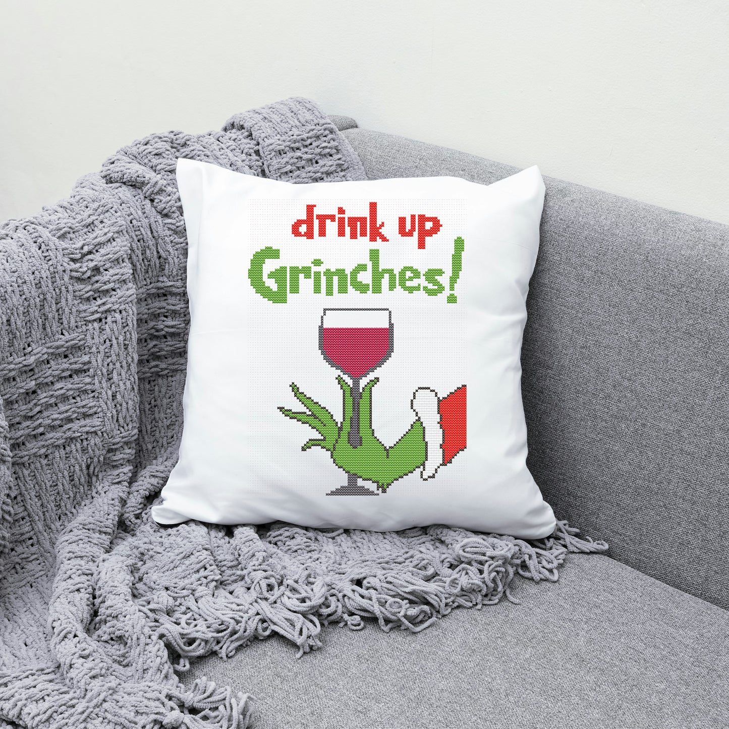 Decorative pillow with 'drink up Grinches!' text and a Grinch holding a wine glass on a gray couch.