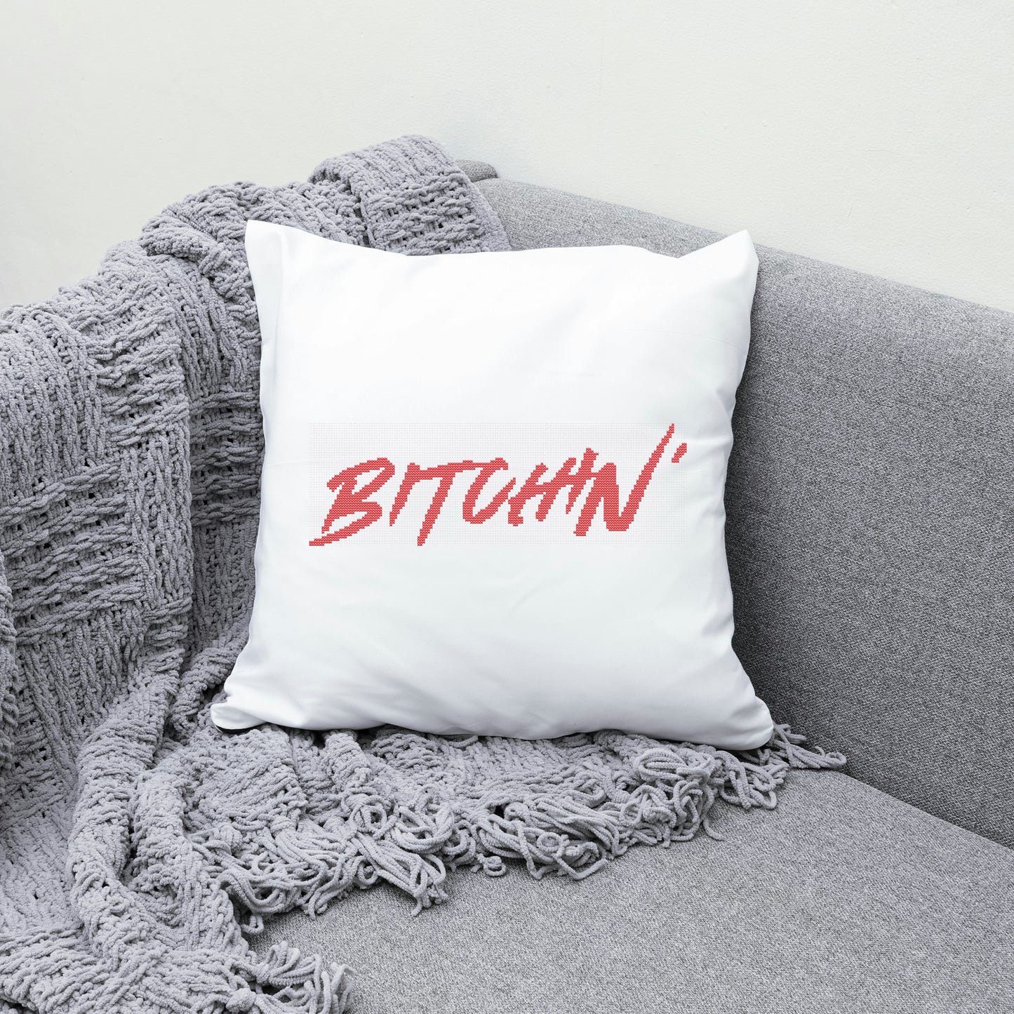 White pillow with 'BITCHIN'' in red on a gray couch with a gray blanket.