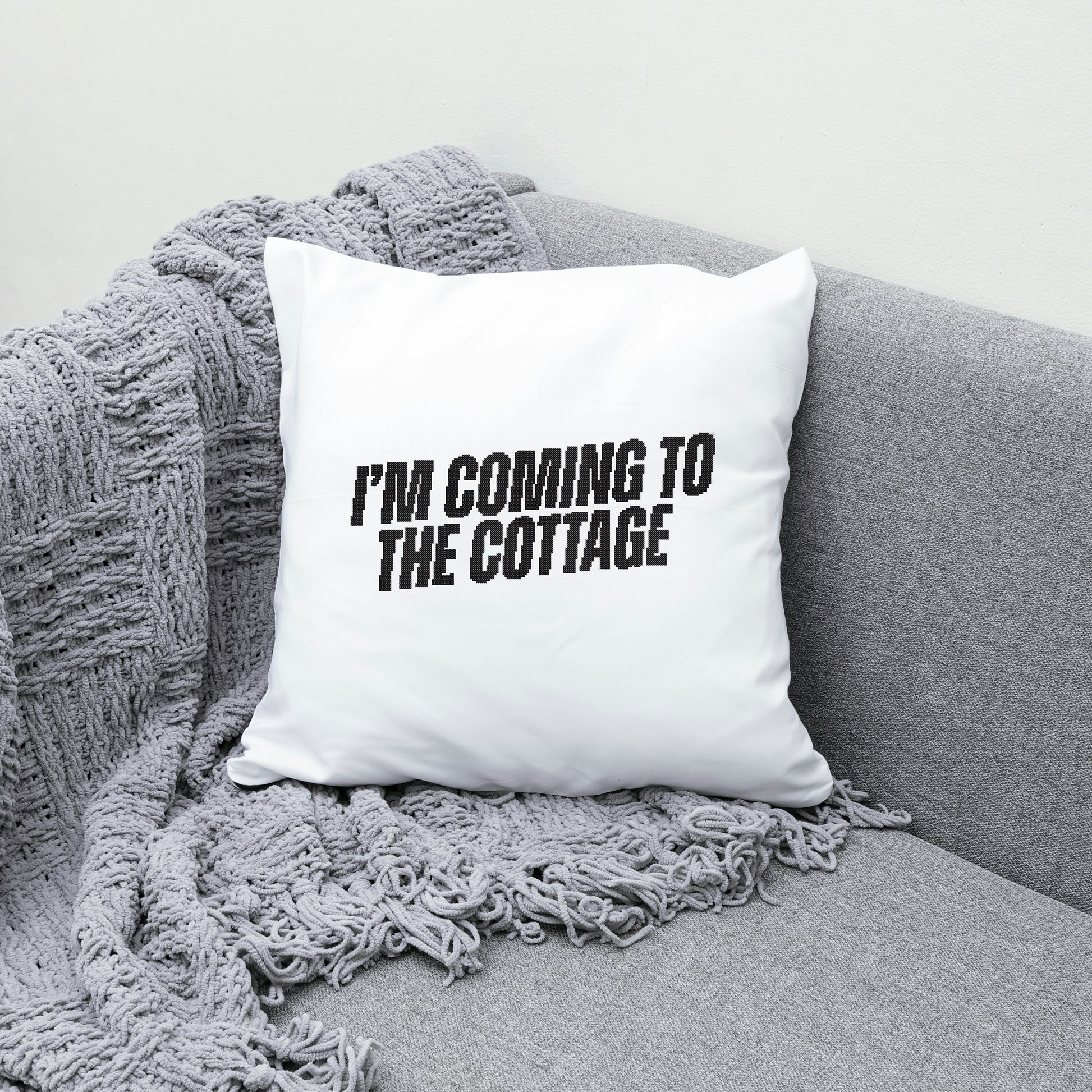 White pillow with 'I'm coming to the cottage' text on a gray couch with a blanket.