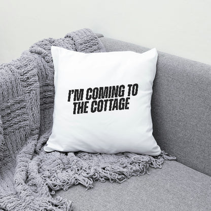 White pillow with 'I'm coming to the cottage' text on a gray couch with a blanket.