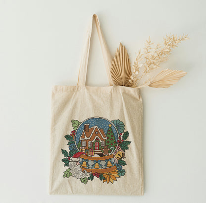 Beige tote bag with a festive design featuring a house and trees, on a light background.