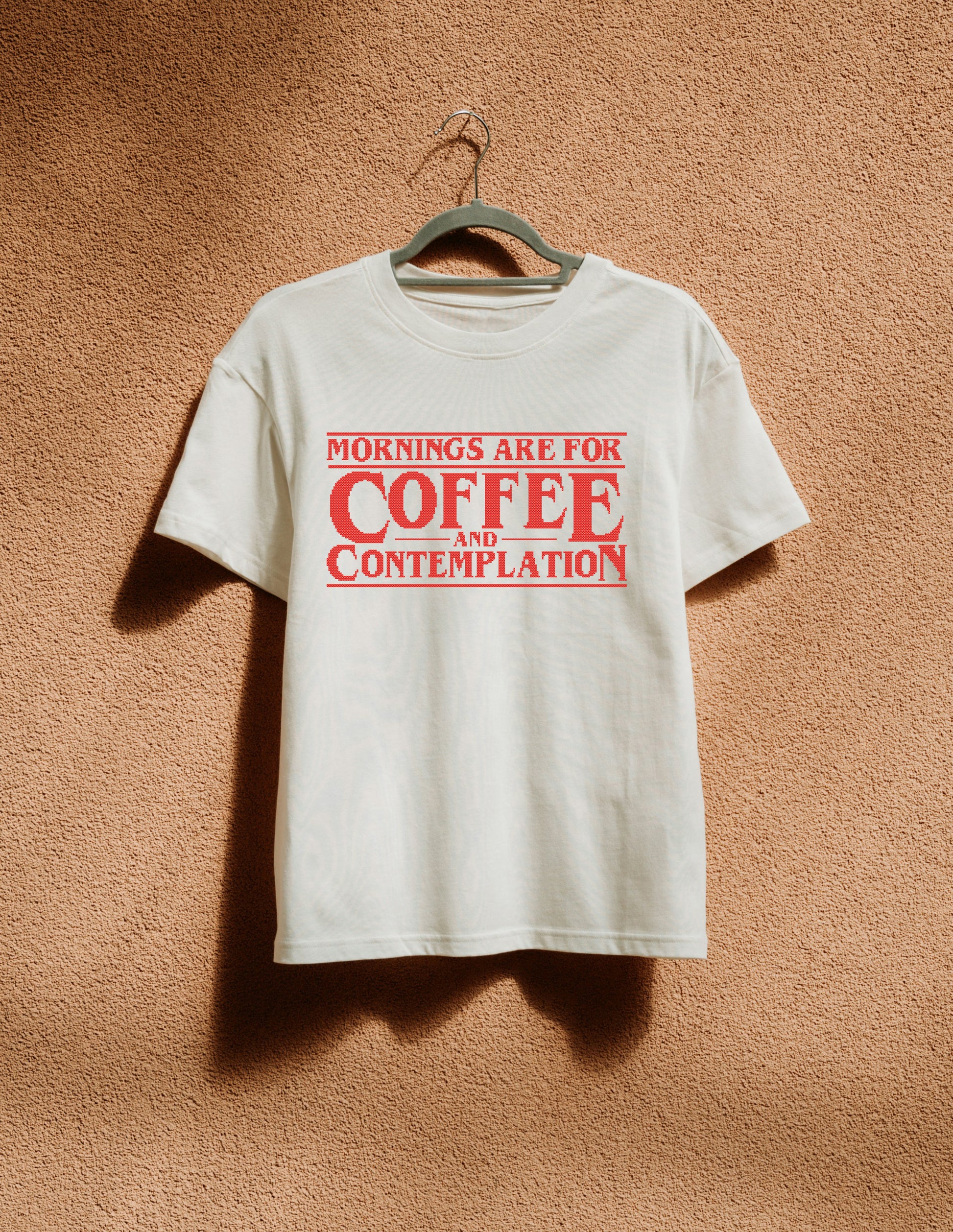 White t-shirt with red text 'Mornings are for Coffee and Contemplation' on a hanger against a brown textured wall.