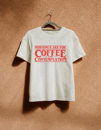White t-shirt with red text 'Mornings are for Coffee and Contemplation' on a hanger against a brown textured wall.