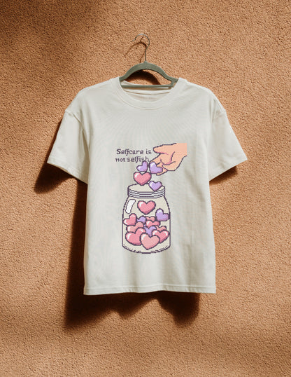 White t-shirt with a graphic design of hearts in a jar and text 'Selfcare is not selfish' on a beige background.