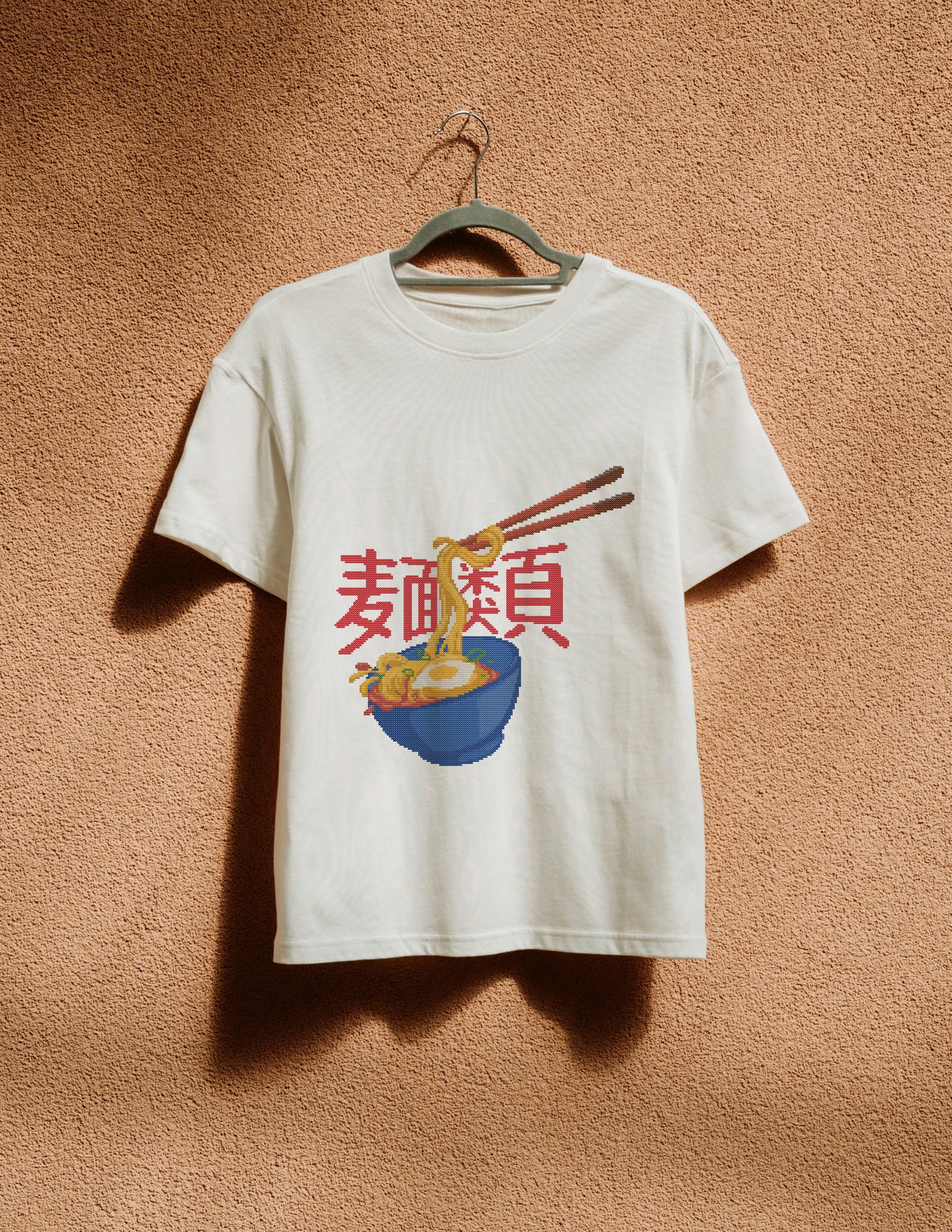 White t-shirt with a graphic design of noodles and chopsticks on a beige wall.