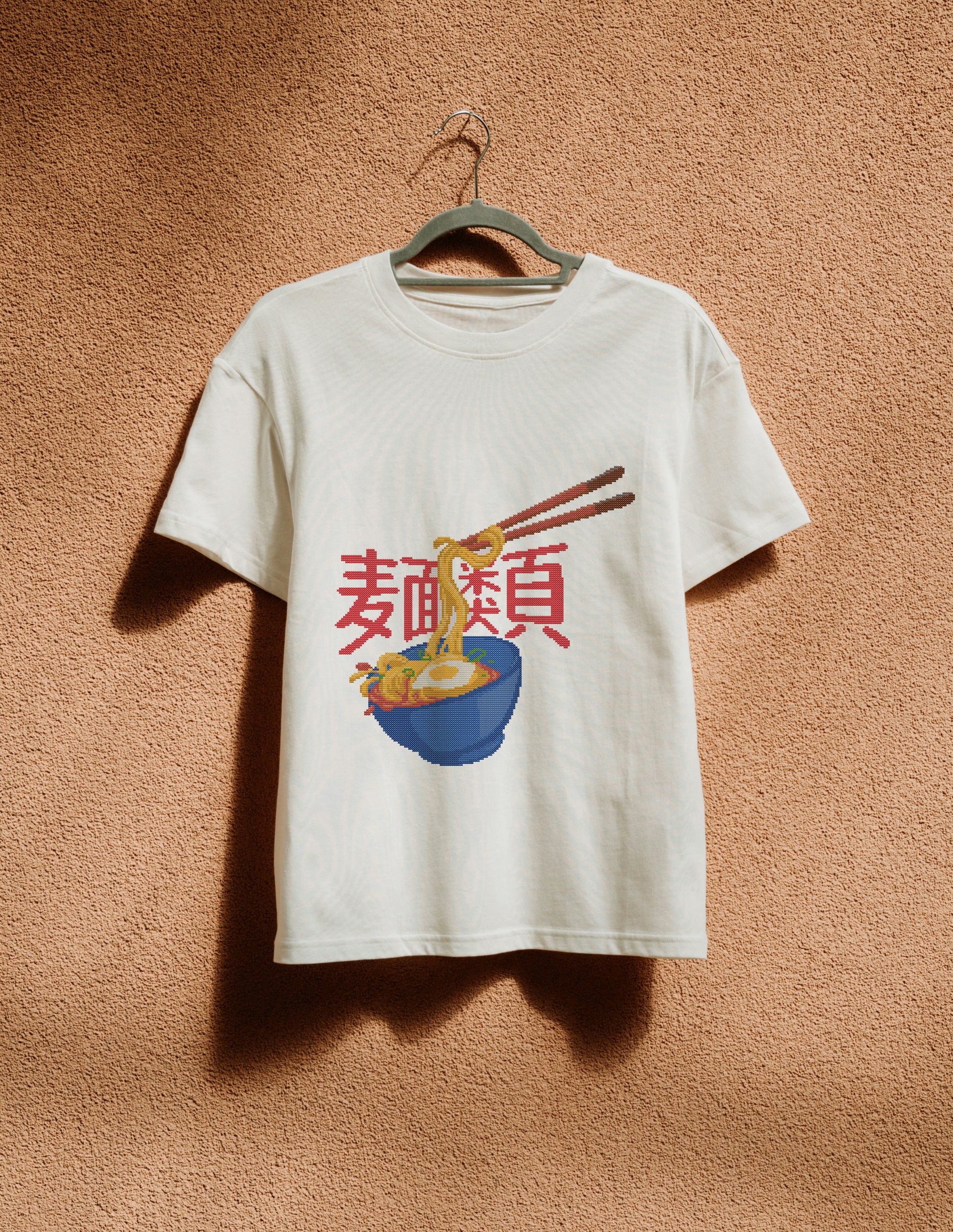 White t-shirt with a graphic design of noodles and chopsticks on a beige wall.