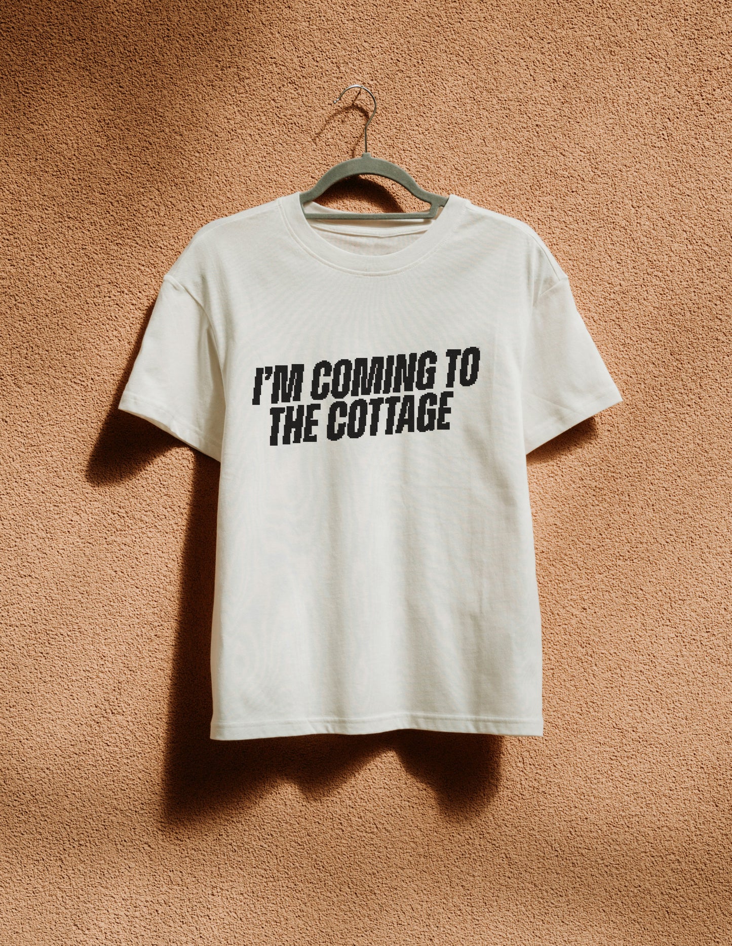 White t-shirt with 'I'm Coming to the Cottage' text on a hanger against a brown textured wall.