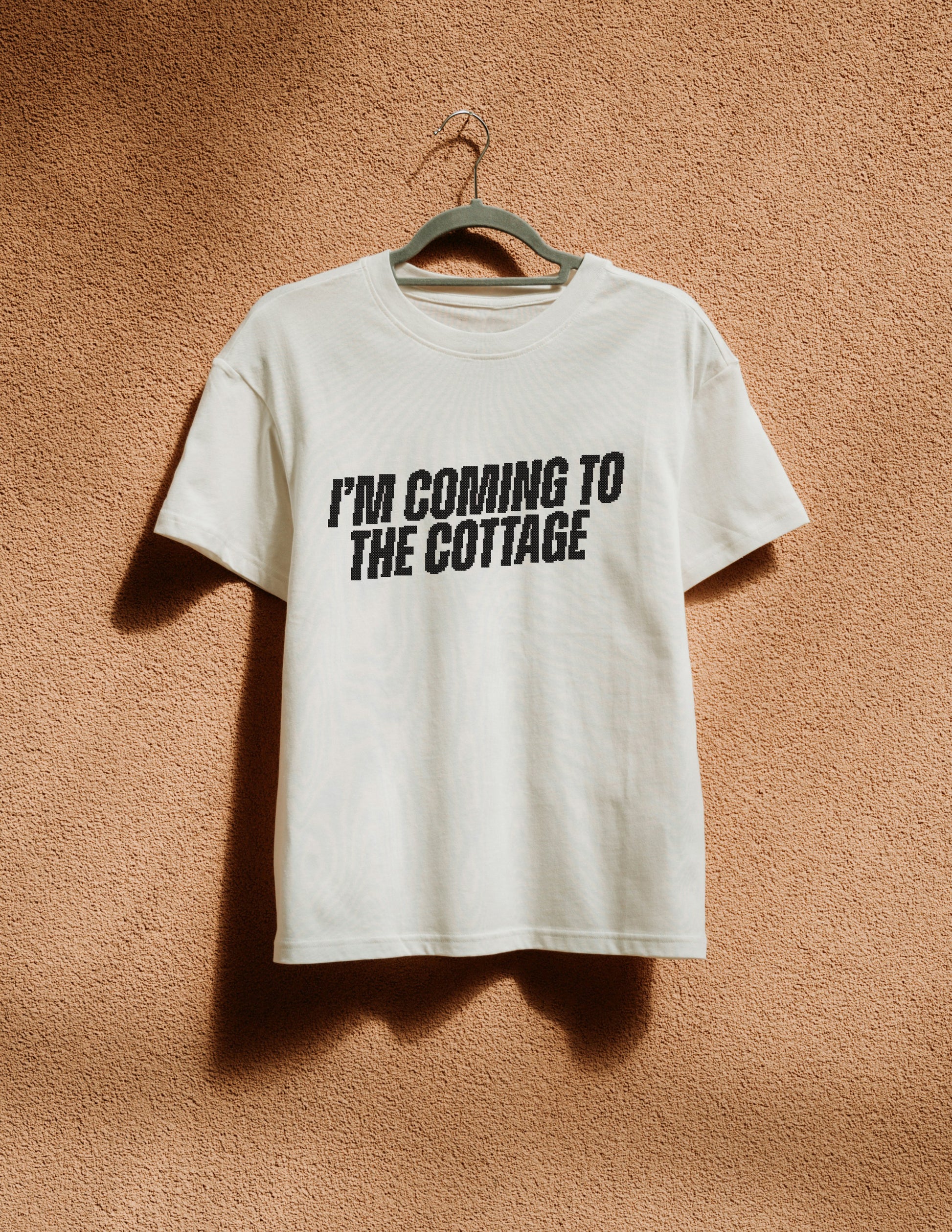 White t-shirt with 'I'm Coming to the Cottage' text on a hanger against a brown textured wall.