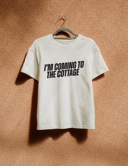 White t-shirt with 'I'm Coming to the Cottage' text on a hanger against a brown textured wall.