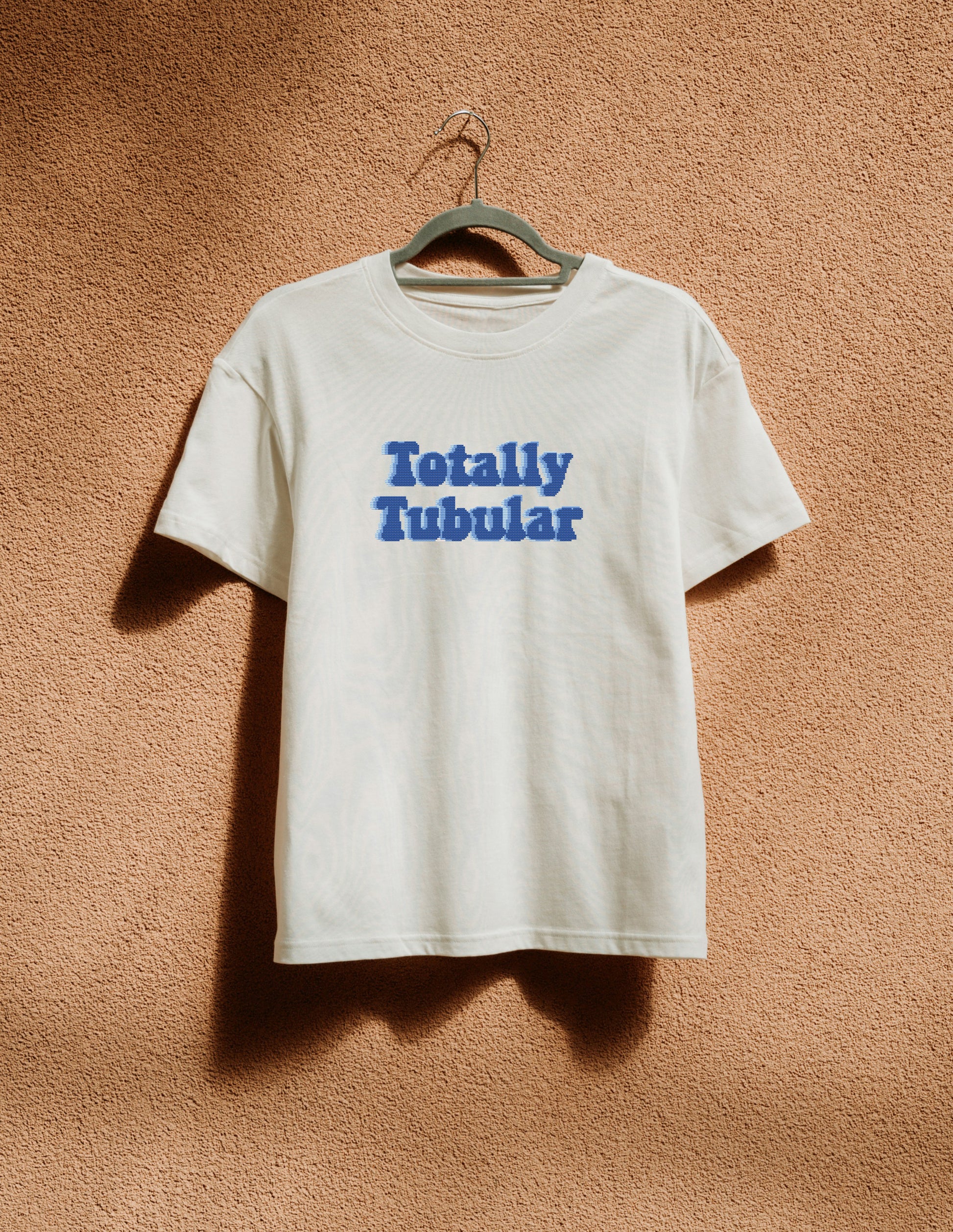 White t-shirt with 'Totally Tubular' text on a hanger against a brown textured wall
