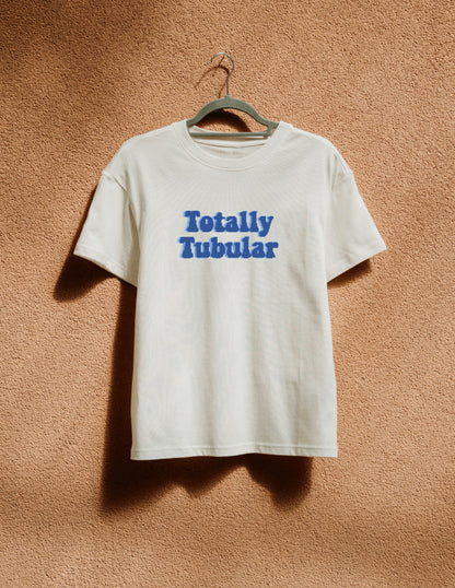 White t-shirt with 'Totally Tubular' text on a hanger against a brown textured wall