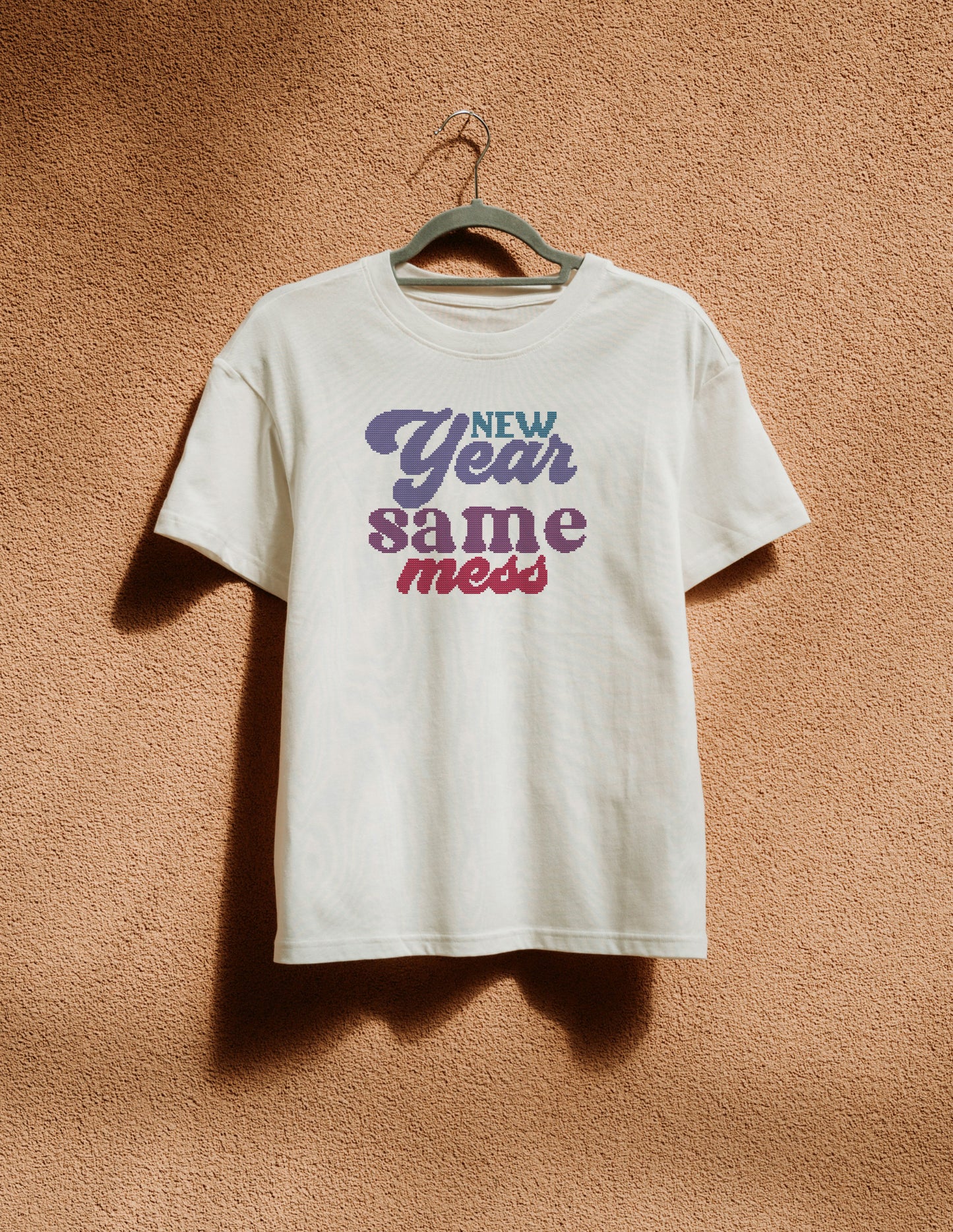 White t-shirt with 'New Year Same Mess' text on a hanger against a brown textured wall.