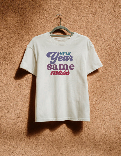 White t-shirt with 'New Year Same Mess' text on a hanger against a brown textured wall.
