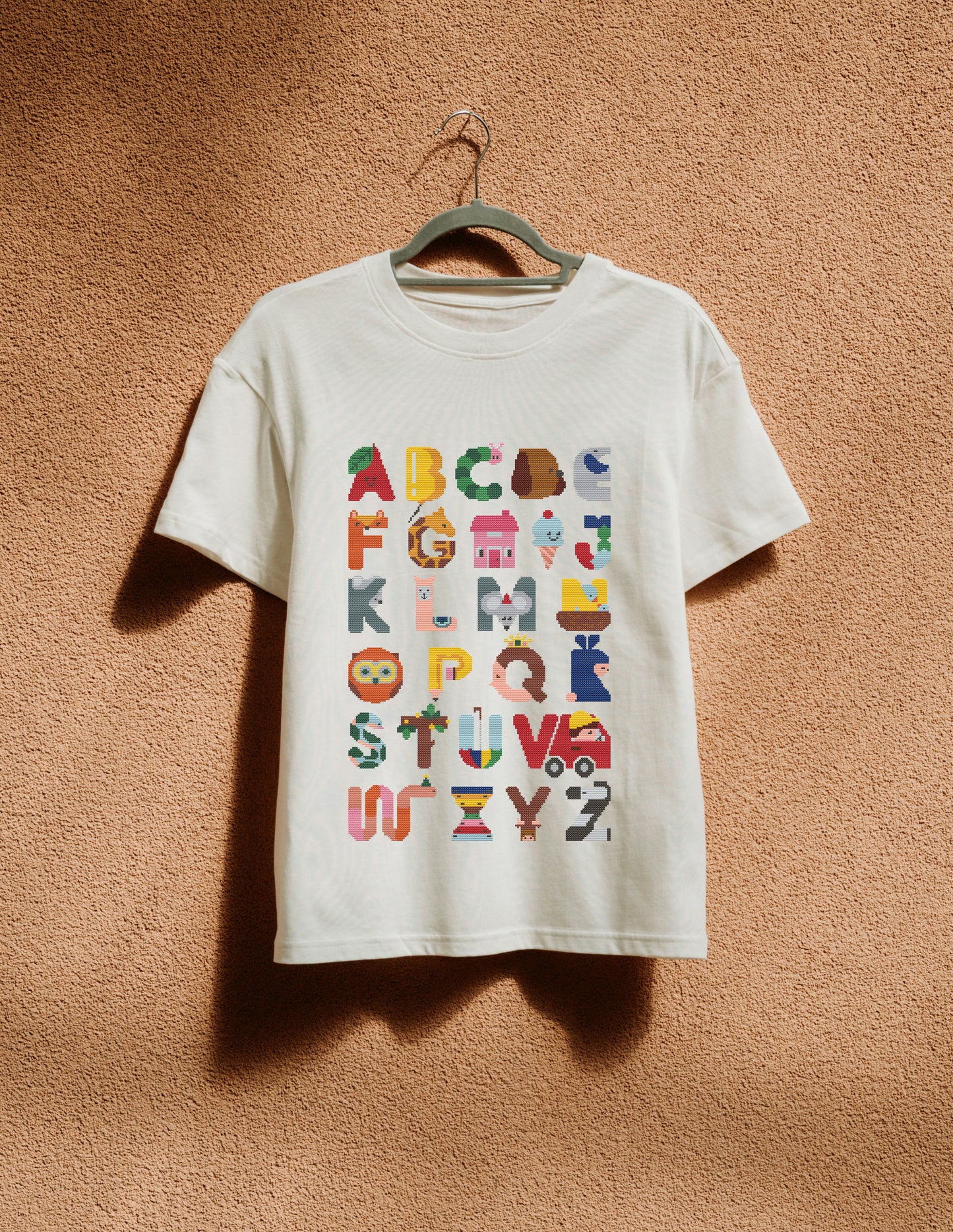 White t-shirt with colorful alphabet design on a hanger against a brown wall.