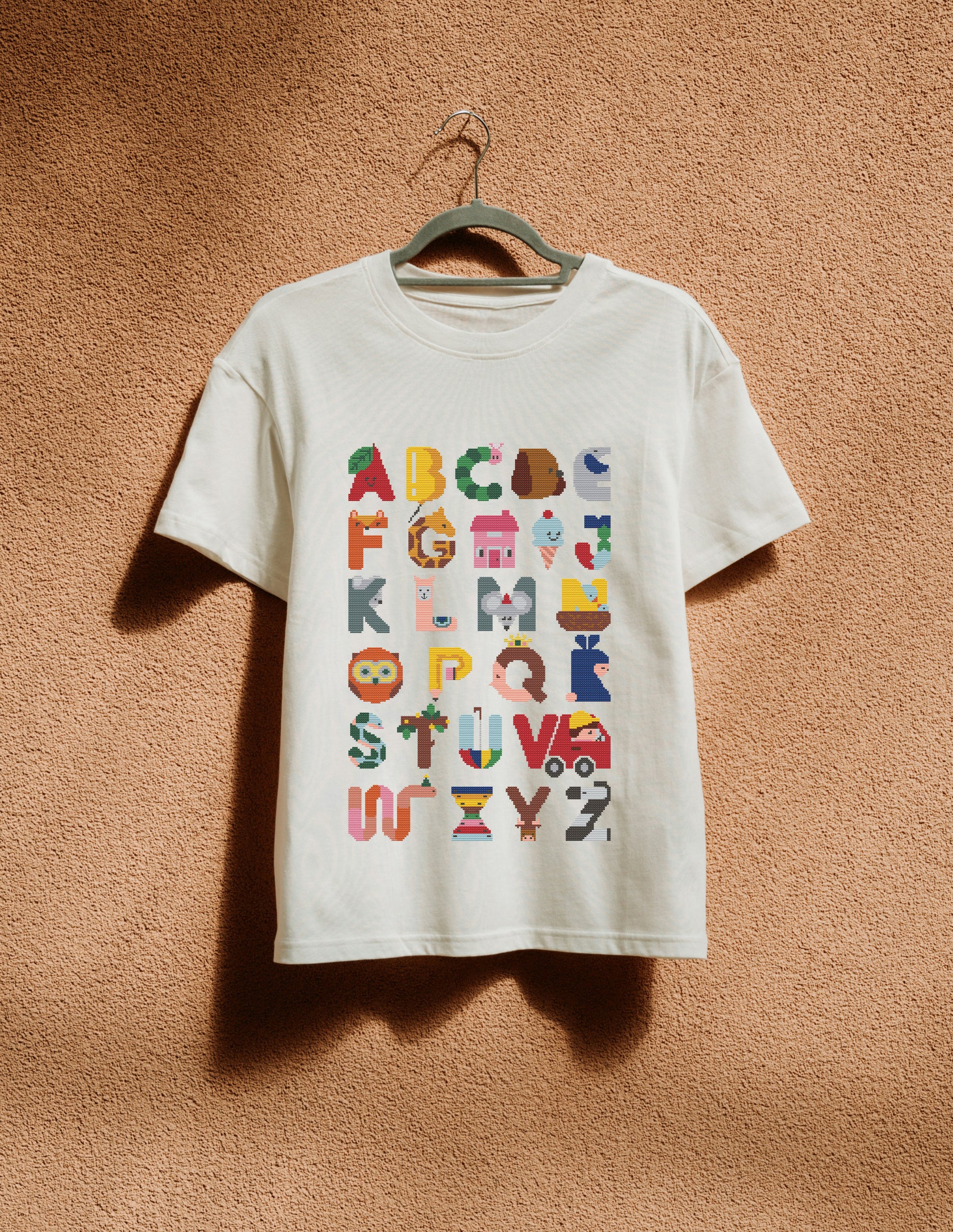 White t-shirt with colorful alphabet design on a hanger against a brown wall.