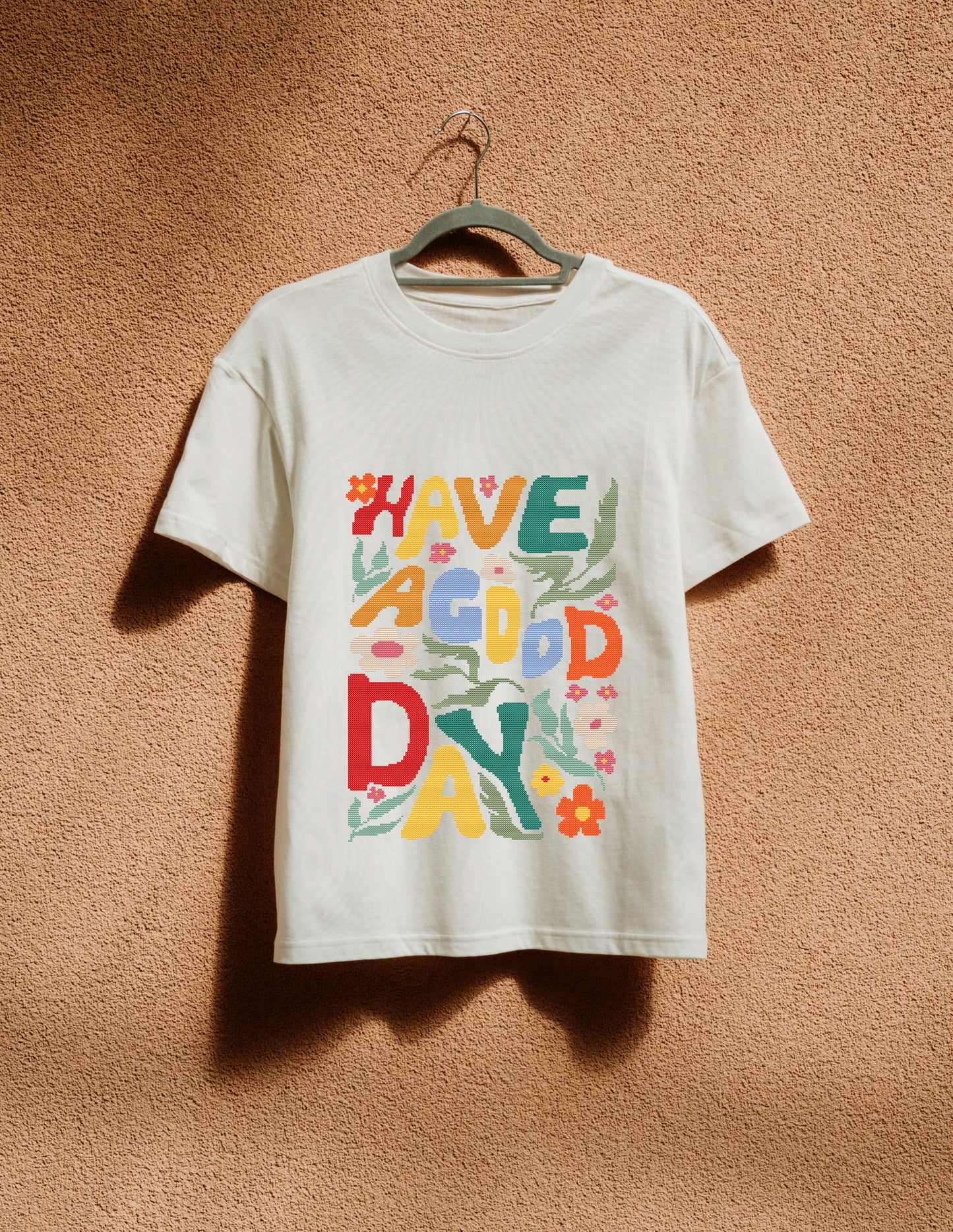 White t-shirt with colorful text 'HAVE A GOOD DAY' on a hanger against a brown textured wall.