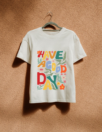 White t-shirt with colorful text 'HAVE A GOOD DAY' on a hanger against a brown textured wall.