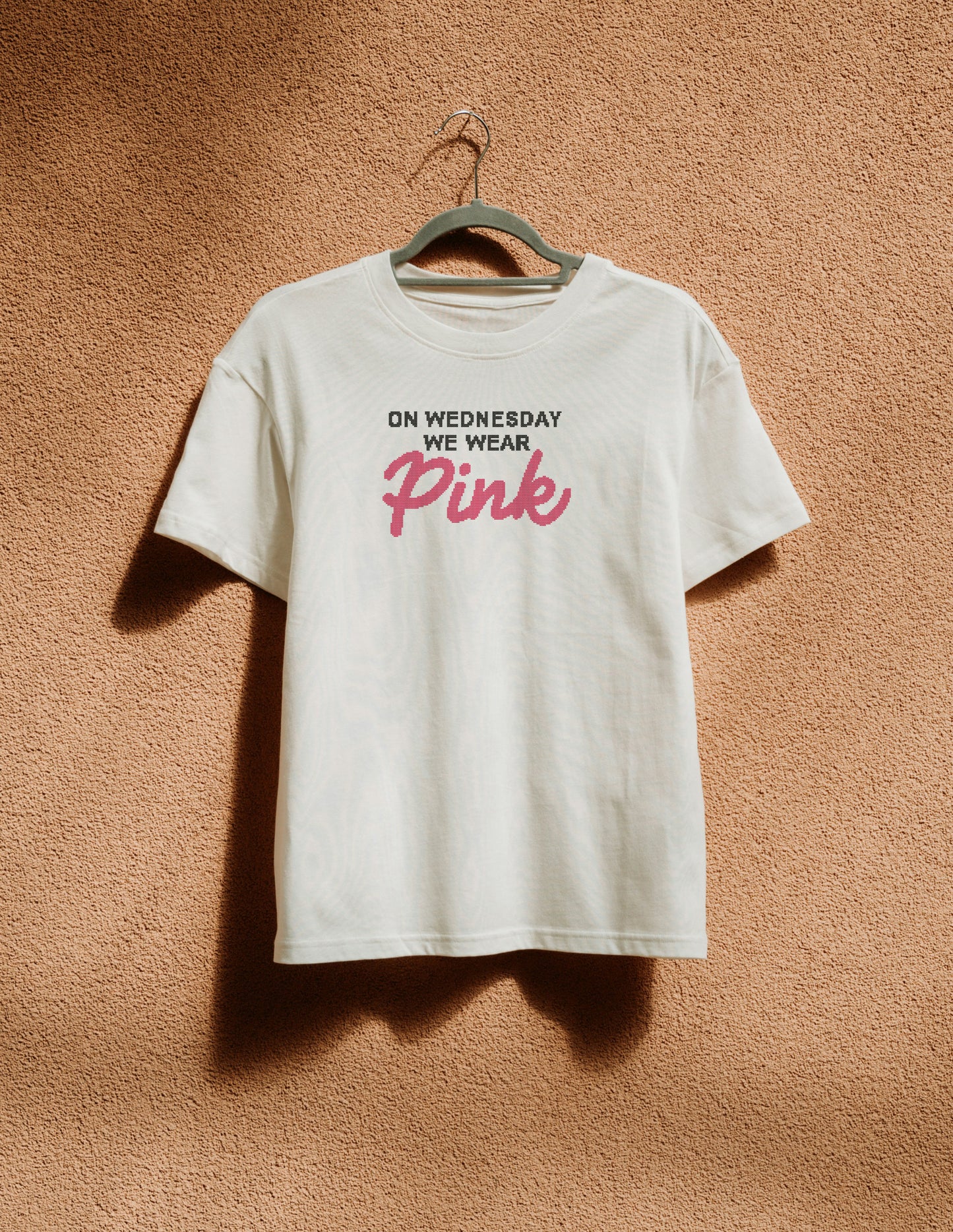 White t-shirt with 'BITCHIN'' in red on a hanger against a brown textured wall.