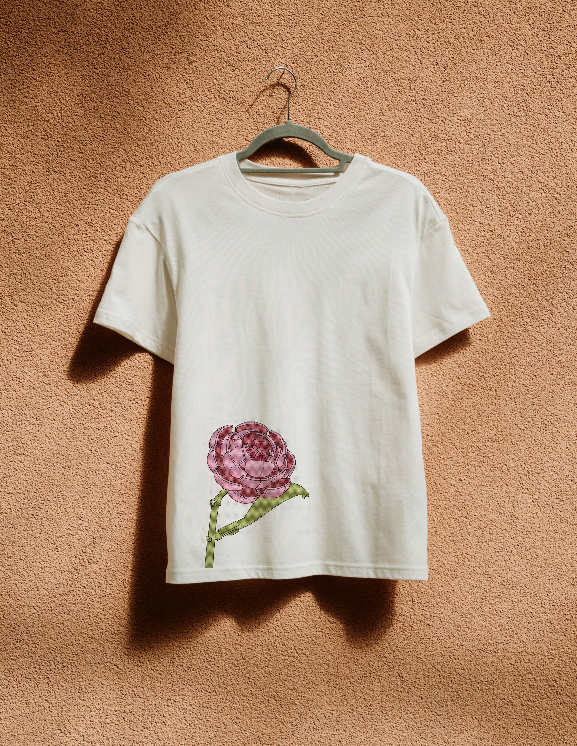White t-shirt with a pink flower design on a hanger against a brown wall.