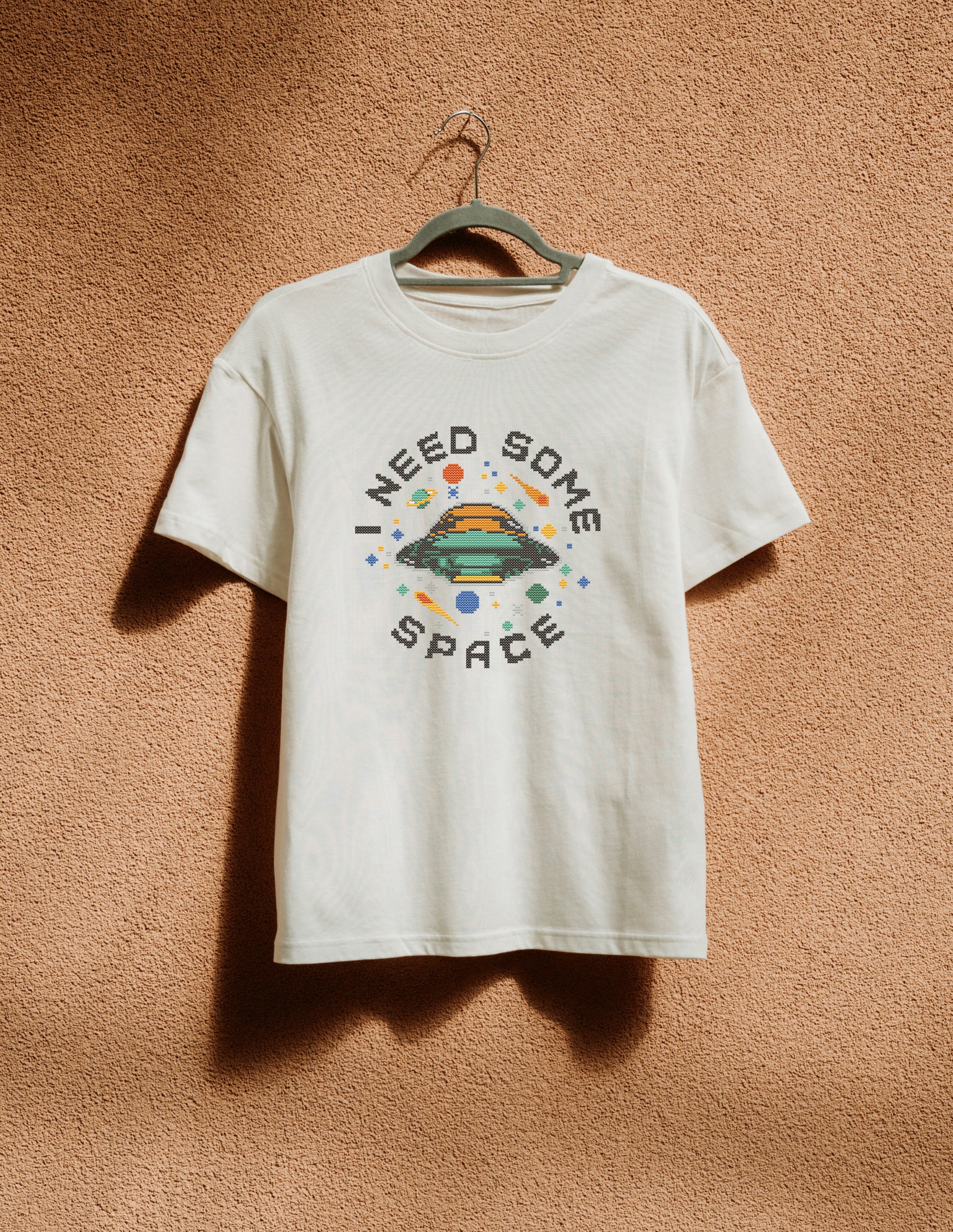 White t-shirt with 'I Need Some Space' graphic on a hanger against a brown textured wall.