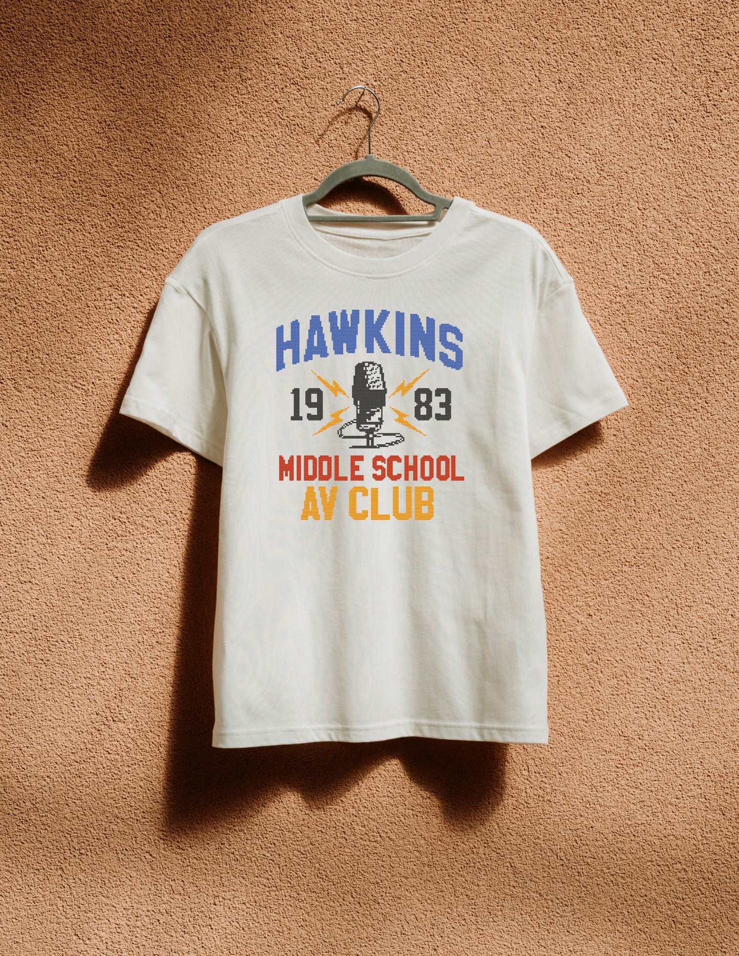 White t-shirt with 'Hawkins Middle School AV Club' design on a hanger against a brown textured wall.