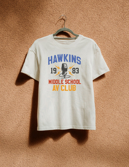 White t-shirt with 'Hawkins Middle School AV Club' design on a hanger against a brown textured wall.