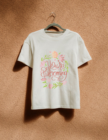 White t-shirt with 'You're Blooming' text and floral design on a hanger against a brown textured wall.