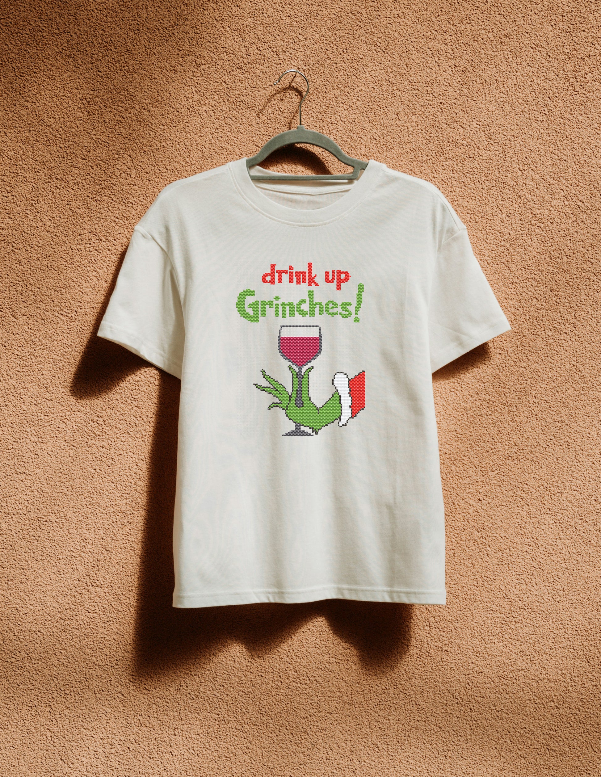 White t-shirt with 'drink up Grinches!' design on a hanger against a brown textured wall.