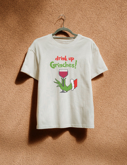 White t-shirt with 'drink up Grinches!' design on a hanger against a brown textured wall.
