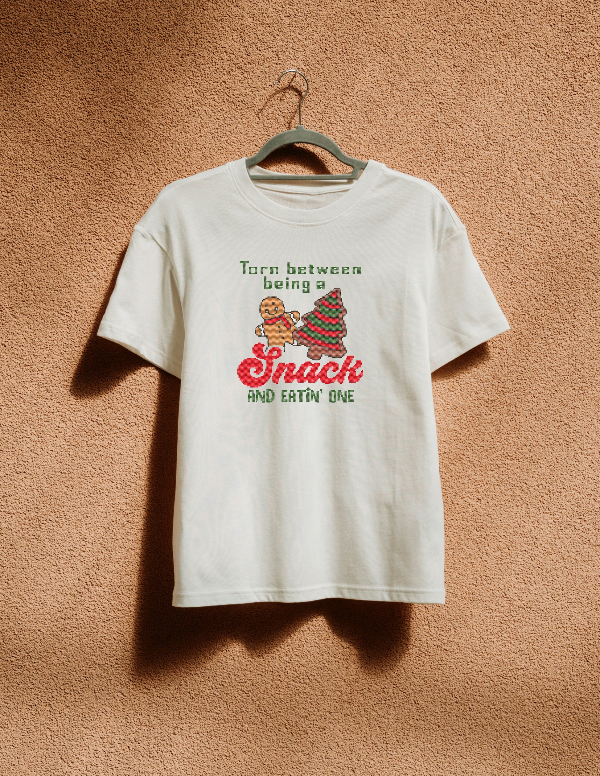 White t-shirt with Christmas-themed design on a hanger against a brown textured wall.