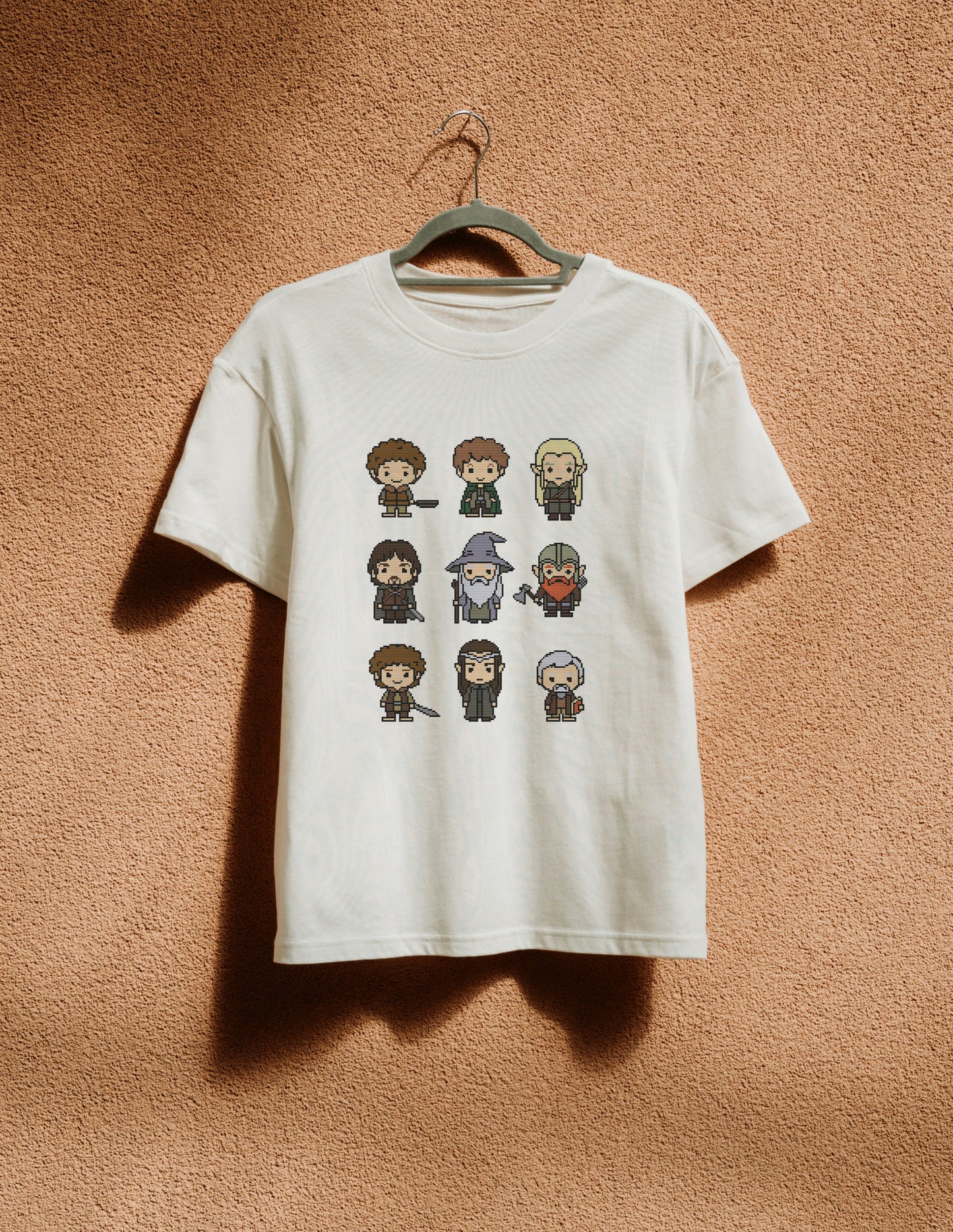 White t-shirt with pixelated character designs hanging on a hanger against a brown textured wall.