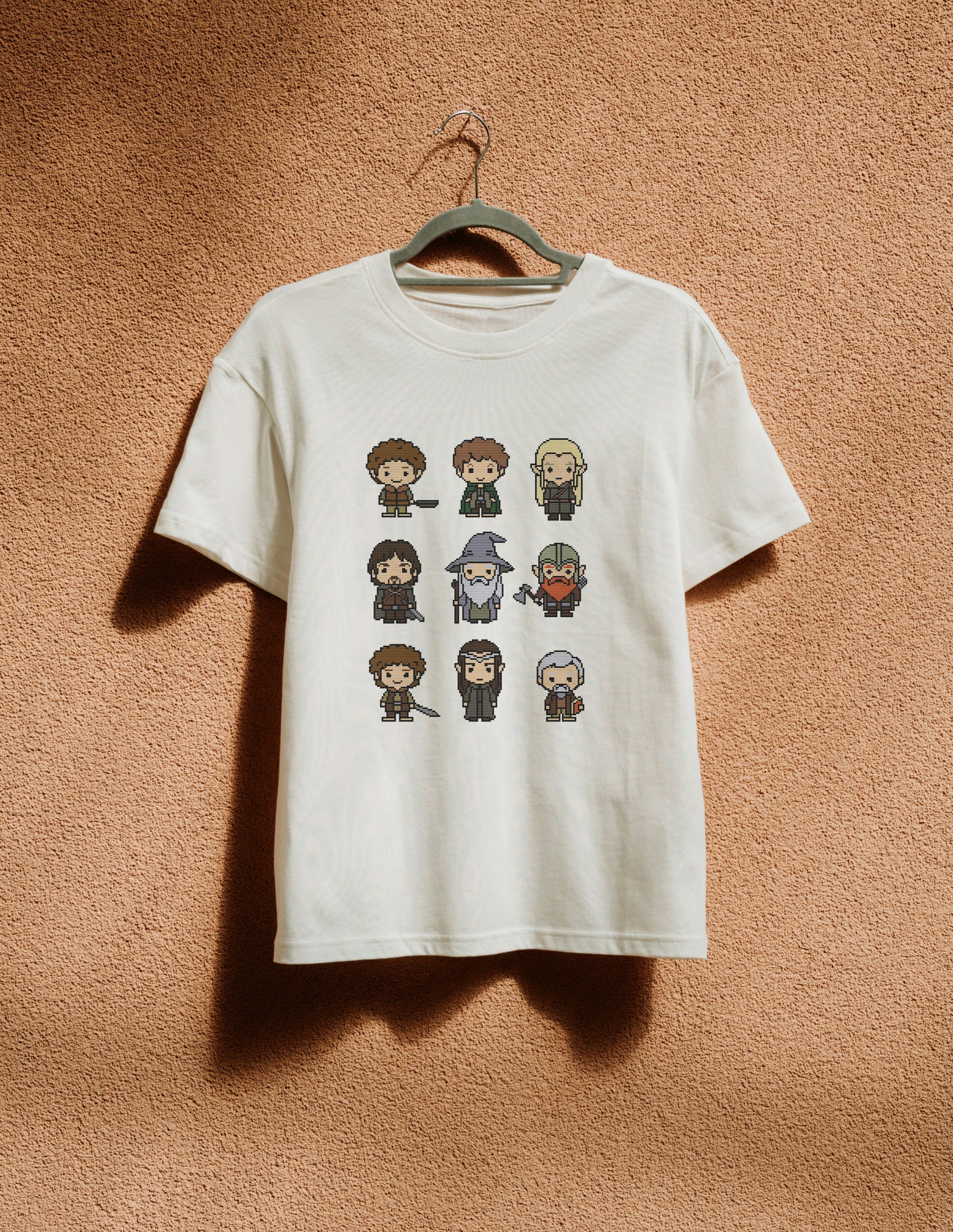 White t-shirt with pixelated character designs hanging on a hanger against a brown textured wall.