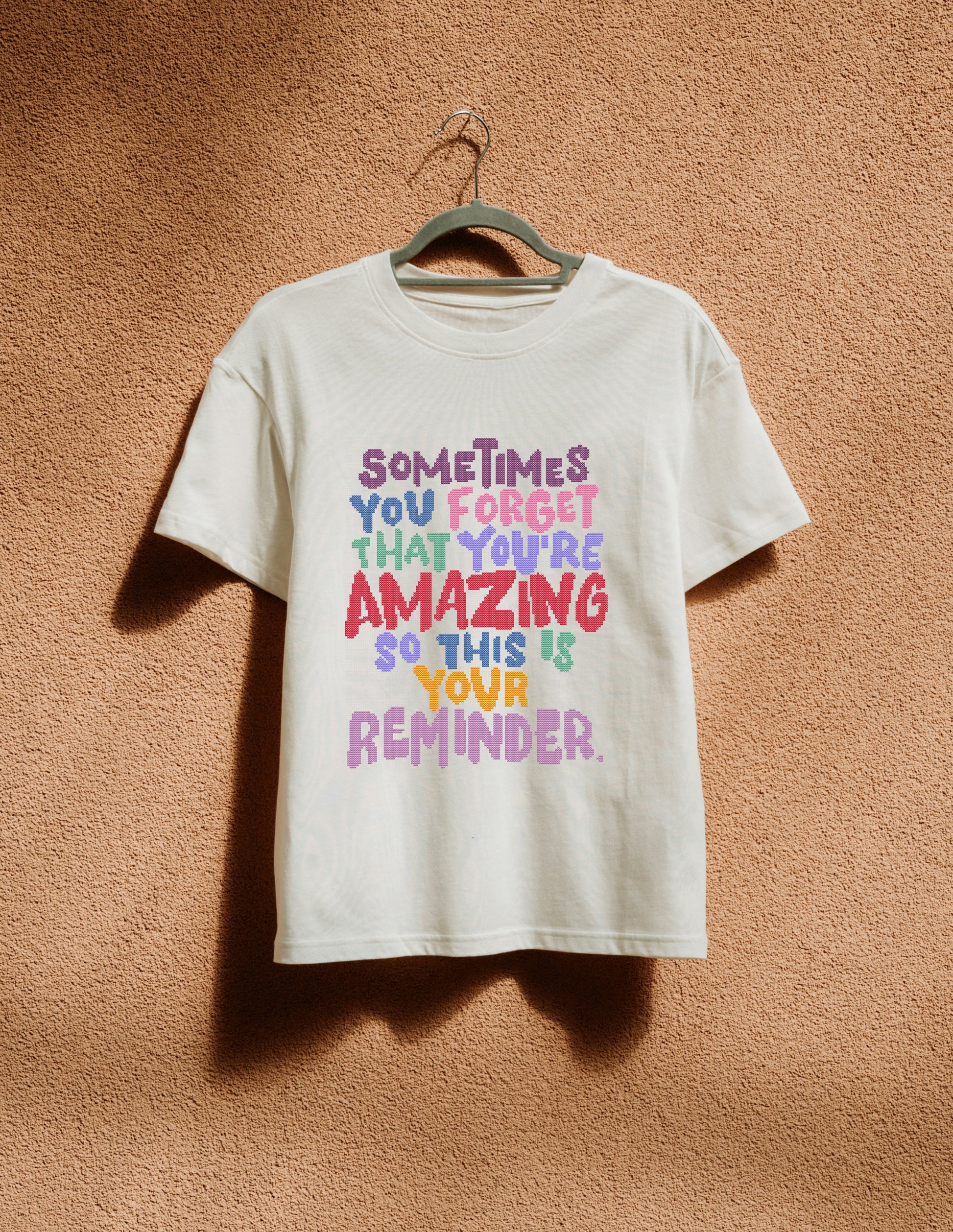 White t-shirt with colorful text on a hanger against a brown textured wall