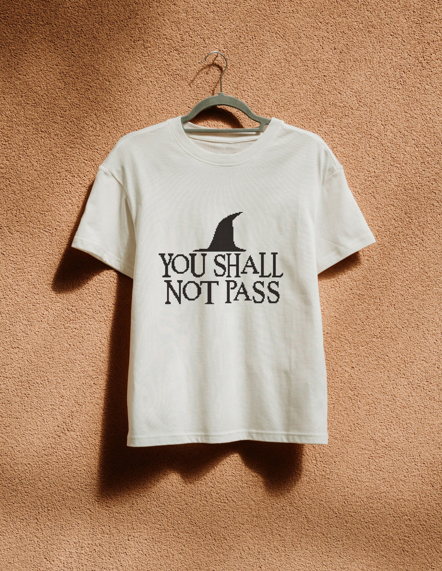 White t-shirt with 'You Shall Not Pass' text and logo on a hanger against a brown background