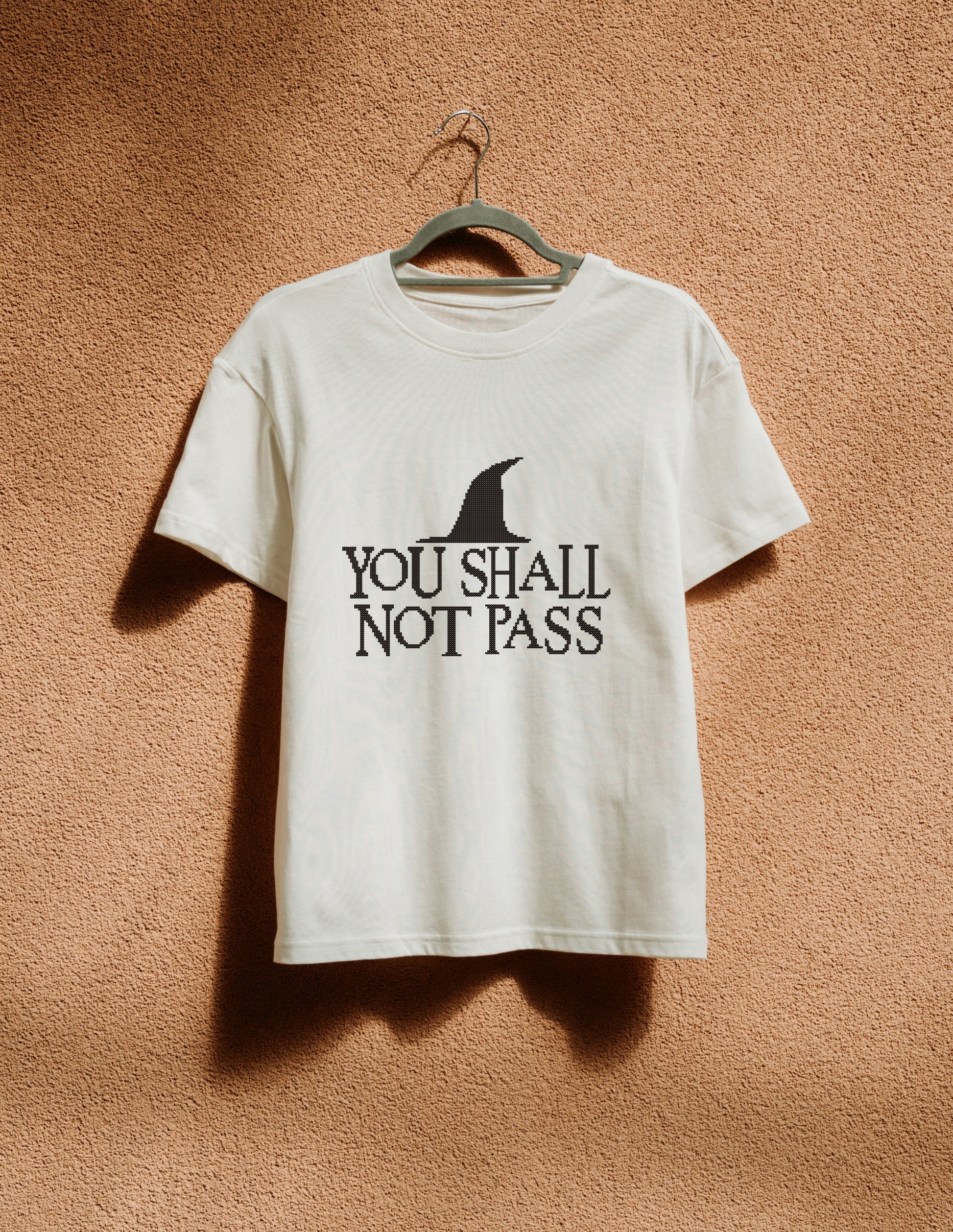 White t-shirt with 'You Shall Not Pass' text and logo on a hanger against a brown background